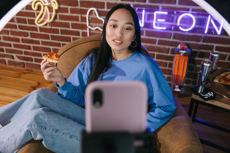 junge Streamerin sitzt vor Handy und Leuchte und hat Pizza Stück in der Hand