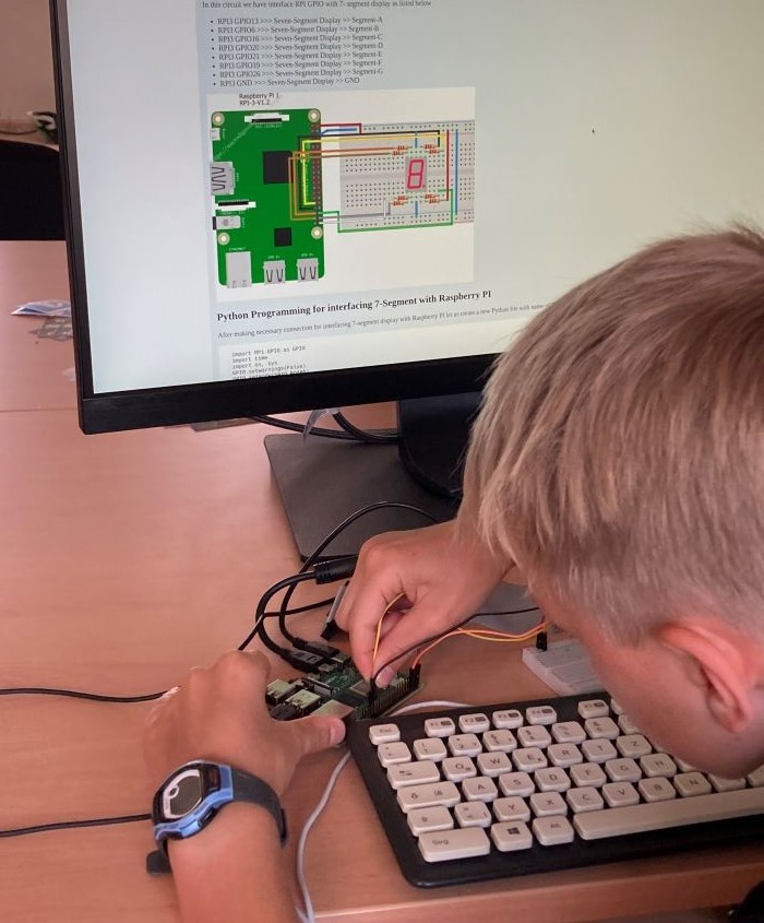Ein Kind steckt einen Draht auf eine Platine. Im Hintergrund ist die Anleitung für einen Mini-Computer.
